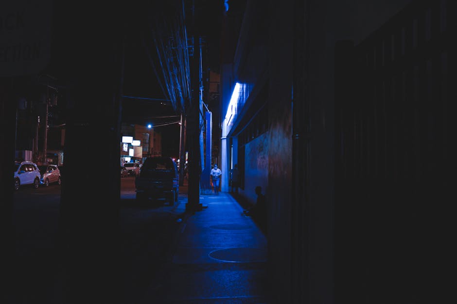 A person walks along a dimly lit street at night, illuminated by neon blue lights.