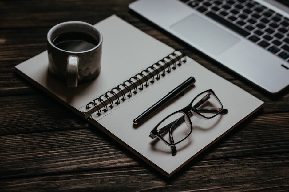 Cozy workspace with a notebook, coffee, glasses, and laptop on a wooden table.