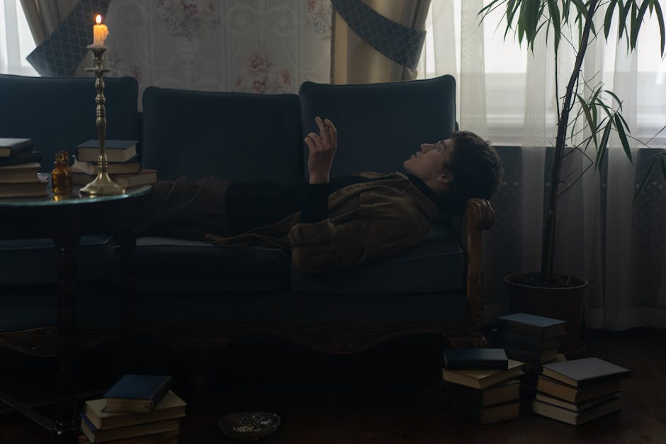 A man lies on a couch in a dimly lit room with candlelight, surrounded by books.
