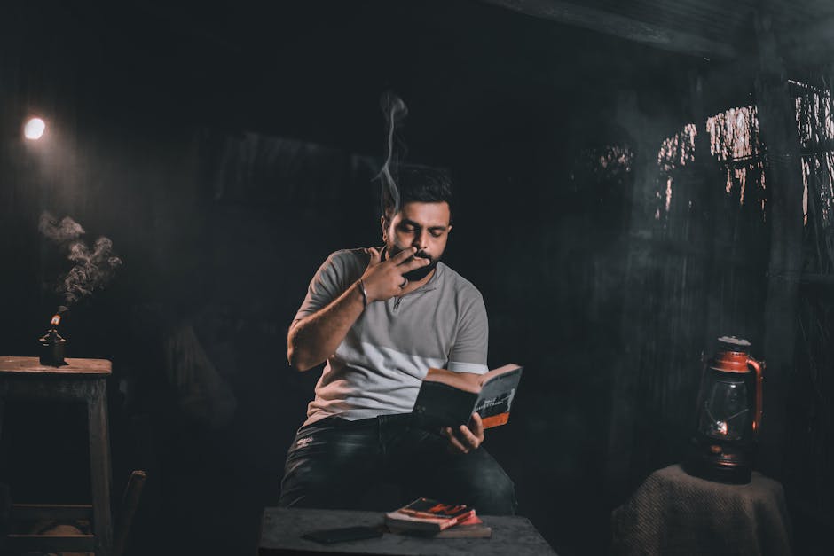 A man sits in a dimly lit room, reading a book and smoking, creating a moody atmosphere.